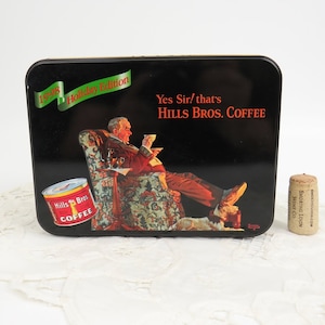 May include: A black metal tin with a 1998 Holiday Edition Hills Bros. Coffee label. The tin features a man sitting in a chair with a cup of coffee and a dog at his feet. The text "Yes Sir/that's Hills Bros. Coffee" is printed on the tin.