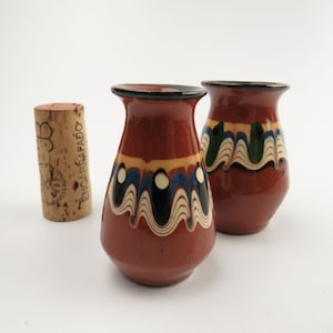 May include: Two small, brown ceramic vases with colorful painted designs. The vases have a rounded body and a narrow neck. The designs feature wavy lines, dots, and swirls in shades of blue, green, white, and black.