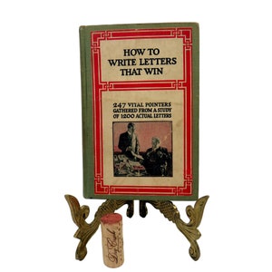 May include: A vintage green book titled "How to Write Letters That Win" with a red border and a picture of two men in suits on the cover. The book is on a gold metal stand.