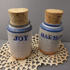 May include: Two ceramic jars with cork lids. The jars are white with blue stripes and blue lettering. One jar says "JOY" and the other says "HARMONY".