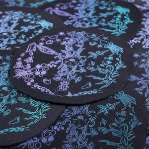 May include: A collection of black circular patches with intricate designs in shades of purple and teal. Each patch features a central figure with flowing hair, surrounded by botanical and celestial elements, including bees and crystals.