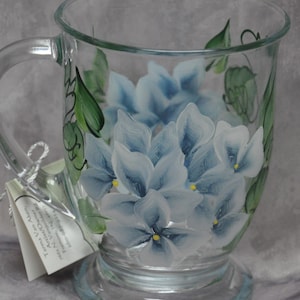 May include: A clear glass mug with a painted design of blue and white flowers with yellow centers. The flowers are painted on the side of the mug.