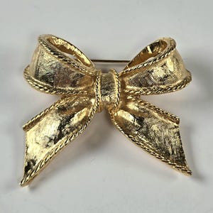 May include: A gold-toned bow brooch with a textured surface and rope-like trim. The bow is tied in the center with a pin on the back for secure fastening. This vintage-style accessory adds a touch of elegance.
