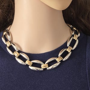 May include: A chunky chain necklace with alternating silver and gold-tone links. The necklace is displayed on a mannequin, showcasing its bold design and contrasting metallic colors. The necklace is a statement piece.