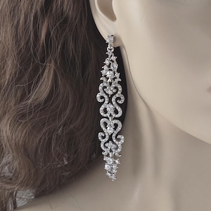 May include: Elegant silver-toned chandelier earrings with a cascading design. The earrings feature a series of sparkling clear crystals arranged in an intricate, flowing pattern. The design includes floral and scroll-like elements, creating a glamorous and sophisticated look.