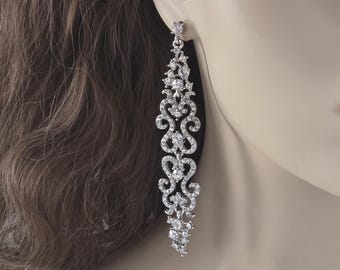 May include: Elegant silver-toned chandelier earrings with a cascading design. The earrings feature a series of sparkling clear crystals arranged in an intricate, flowing pattern. The design includes floral and scroll-like elements, creating a glamorous and sophisticated look.