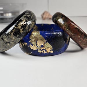 May include: Three decorative bangles and a small bowl. The bangles are black, red, and black with gold flecks. The bowl is blue with gold leaf accents. The items are on a white surface.