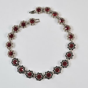 May include: A silver-toned necklace with a floral design. The necklace features alternating red and black gemstone clusters. The red gemstones are surrounded by silver-toned metal, and the black gemstones are in a floral pattern.