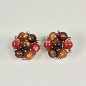 May include: A pair of stud earrings featuring a cluster of round beads in shades of red, brown, and gold. The beads are arranged in a floral pattern and attached to silver-colored posts. The earrings are set against a white background.