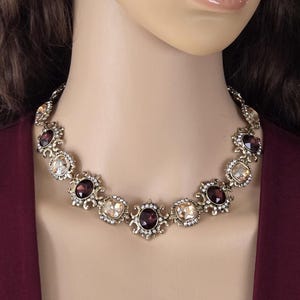 May include: A gold-toned necklace featuring alternating oval and square-cut gemstones in shades of burgundy and champagne. The gemstones are set in ornate, filigree-style settings, creating a vintage-inspired look. The necklace is displayed on a mannequin.