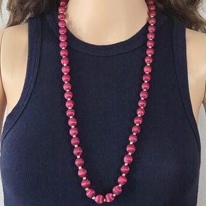 May include: A long, magenta beaded necklace with gold-colored spacers. The necklace is draped over a dark blue tank top. The beads appear to be round and evenly spaced, creating a classic and elegant look.