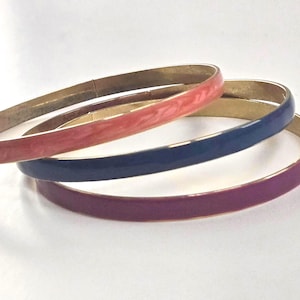 May include: Three colorful bangles in shades of coral, navy blue, and purple. The bracelets have a gold-tone metal base and are stacked on a white surface. These accessories are perfect for adding a pop of color to any outfit.