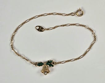 May include: Gold-toned bracelet featuring a Christmas bell charm. The bell is adorned with green enamel holly leaves and a clear crystal. The bracelet has a delicate chain and a secure clasp, perfect for festive occasions.