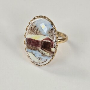 May include: Gold-toned ring featuring an oval-shaped, hand-painted miniature landscape scene. The artwork depicts a red building in a snowy environment, framed by a decorative gold border. The ring's band is a simple, polished gold.