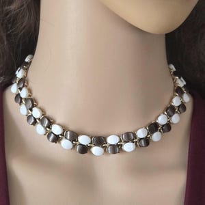May include: A vintage necklace featuring a double strand design. The necklace alternates between white and brown oval-shaped beads, set in a gold-tone metal frame. The necklace is a choker style.