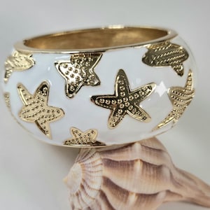 May include: A white and gold bangle bracelet featuring gold starfish and seashell designs. The bracelet has a gold interior and is resting on a seashell. The bracelet is a beach-themed accessory.
