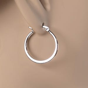 May include: A close-up of a silver hoop earring. The earring is round and has a shiny, metallic finish. The earring is attached to an ear. The background is a neutral beige color.