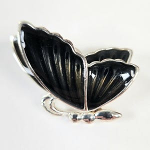 May include: A silver-toned butterfly brooch with black enamel wings. The brooch has a detailed design with a shiny finish. The butterfly's body is made of silver, and the wings are black with a textured pattern.