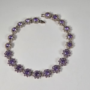 May include: A silver-toned necklace featuring a series of purple gemstone flower-shaped clusters. Each cluster is composed of a central, larger purple stone surrounded by smaller, matching stones. The necklace is designed with a clasp closure.