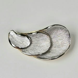 May include: A silver-toned brooch with a layered, abstract design. The brooch features three overlapping, textured oval shapes with a brushed metal finish. The edges are smooth and polished, creating a contrast with the textured surfaces.
