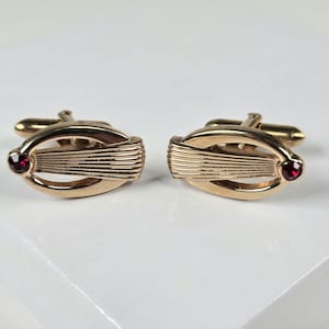 May include: A pair of gold-toned cufflinks. Each cufflink features an oval design with a series of parallel lines and a small, round, red gemstone. The cufflinks have a classic, elegant appearance.