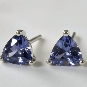 May include: A pair of stud earrings featuring triangular, faceted, blue gemstones set in silver-colored metal. The earrings have a post and a small, round accent at the top and bottom corners of the triangle.