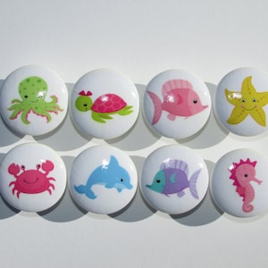 May include: Eight ceramic knobs with different sea creatures. The knobs are white with colorful sea creatures including a pink crab, a green octopus, a blue dolphin, a pink fish, a purple fish, a green turtle, a yellow starfish, and a pink seahorse.