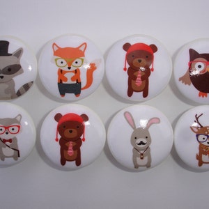 May include: Set of eight round ceramic knobs featuring whimsical animal illustrations. Each knob depicts a different animal wearing glasses and a bowtie, including a raccoon, fox, bear, owl, squirrel, bunny, deer, and another bear.