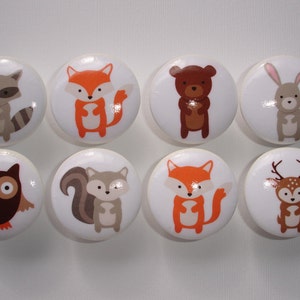 May include: Set of eight ceramic knobs featuring woodland animal illustrations. The knobs are white with colorful animal designs, including a raccoon, fox, bear, rabbit, owl, squirrel, deer, and another fox.