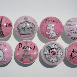 May include: Set of eight pink and white ceramic drawer knobs with various Parisian-themed designs, including the Eiffel Tower, a crown, a poodle, a bicycle, a chandelier, and a floral motif. The knobs have a distressed look and are perfect for adding a touch of Parisian charm to any room.