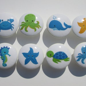 May include: Set of eight ceramic drawer knobs featuring various sea creatures, including a crab, octopus, whale, starfish, seahorse, and turtle. The knobs are white with colorful cartoon illustrations.