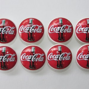 May include: Eight round white ceramic buttons with a red background and a Coca-Cola bottle graphic. The bottle is brown with a white label and white text that reads "Coca-Cola".