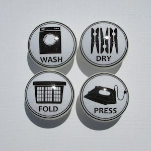 May include: Four white ceramic knobs with black outlines and black icons. The icons depict a washing machine, clothes pins, a laundry basket, and an iron. The knobs are labeled "WASH", "DRY", "FOLD", and "PRESS".