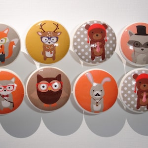 May include: Set of eight round ceramic knobs with various woodland animal illustrations. Each knob features a different animal, including a fox, deer, bear, raccoon, owl, squirrel, rabbit, and a bear wearing a hat. The knobs are decorated with orange, brown, gray, and white colors.