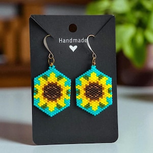 May include: A pair of hexagonal beaded earrings featuring a sunflower design with a brown centre, yellow petals, and a bright turquoise background. The earrings hang from silver-toned metal hooks and are displayed on a black card with the text Handmade.