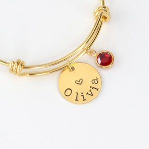 May include: Gold-tone adjustable bangle bracelet with a round charm engraved with the name "Olivia" and a small heart. A red gemstone is suspended from the bracelet. The bracelet is on a white background.