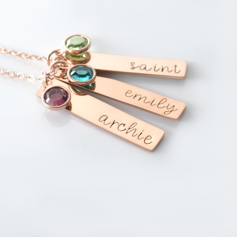 K&ouml;nnte beinhalten: Eine Halskette aus Ros&eacute;gold mit drei personalisierten Bar-Anh&auml;ngern. Die Anh&auml;nger sind mit den Namen "archie", "emily" und "saint" graviert. An jedem Anh&auml;nger ist ein kleiner Edelstein befestigt: ein lila Stein, ein blauer Stein und ein gr&uuml;ner Stein.