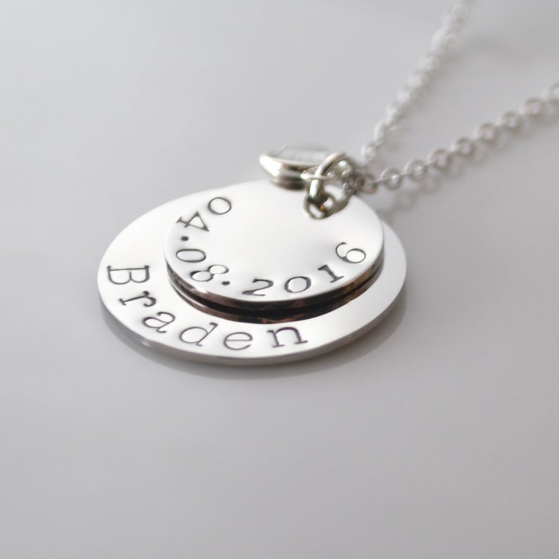 May include: Silver necklace with two round pendants. The top pendant has the date "08.2016" engraved on it. The bottom pendant has the name "Braden" engraved on it.