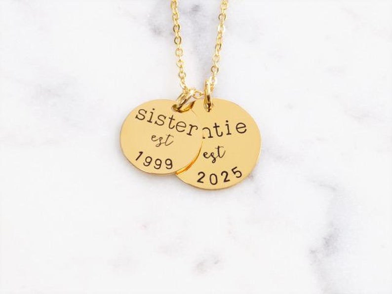 Puede incluir: Collar de oro con dos colgantes redondos. El colgante m&aacute;s grande dice "sistertie est 2025" y el colgante m&aacute;s peque&ntilde;o dice "est 1999".