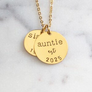 Puede incluir: Collar de oro con dos colgantes redondos. El colgante m&aacute;s grande dice "auntie est 2025" y el colgante m&aacute;s peque&ntilde;o dice "sis".