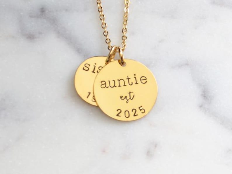 Puede incluir: Collar de oro con dos colgantes redondos. El colgante m&aacute;s grande dice "auntie est 2025" y el colgante m&aacute;s peque&ntilde;o dice "sis".