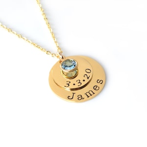 May include: Gold necklace with a layered pendant featuring the name "James" and the date "3.3.20" engraved on two circular discs. A small light blue gemstone is centered on the top disc.