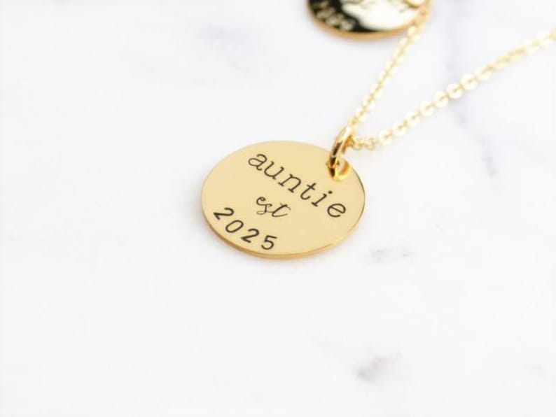 Puede incluir: Collar de oro con un colgante redondo que dice "auntie est 2025".