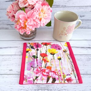 May include: A pink and white floral print fabric coaster with a pink border. The coaster is on a white wooden surface. A mug with a floral design is sitting on the coaster.