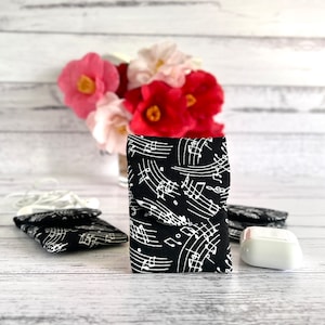 Peut inclure: Un étui en tissu noir et blanc avec un motif de notes de musique. L'étui est rectangulaire et plié. D'autres étuis et des écouteurs blancs sont visibles. Un bouquet de fleurs roses et rouges est en arrière-plan.