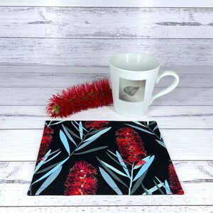 May include: A white ceramic mug with a leaf design, a red bottlebrush flower, and a black coaster with a floral pattern featuring red flowers and blue-grey leaves. The coaster is rectangular.