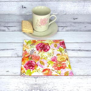 May include: A square floral coaster with a pink border. A teacup and saucer with a floral design and a biscuit are on a white wooden surface. The coaster features red, pink, and orange flowers.