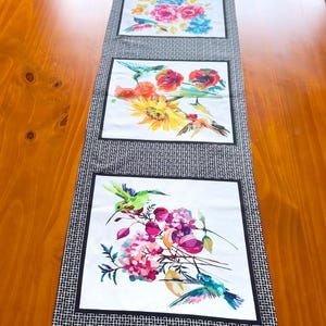 May include: A black and white table runner with three panels featuring colourful watercolour paintings of hummingbirds and flowers. The panels are separated by a black and white chequered pattern.