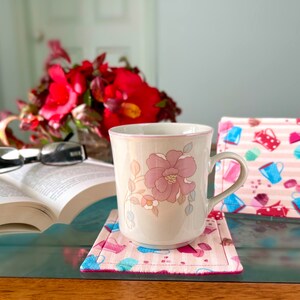May include: A white ceramic mug with a floral design sits on a pink and white patterned coaster. A book and glasses are on the table in the background.