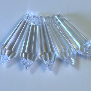May include: Five clear, faceted glass chandelier crystals arranged in a fan shape. Each piece features a pointed, tapered end and a flat, drilled top section for hanging. The transparent glass prisms reflect light, creating sharp, geometric edges.